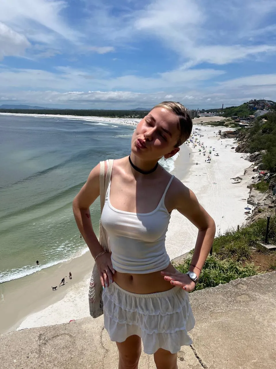 A college girl in a white top and skirt stands on the beach, enjoying the sun and ocean waves.