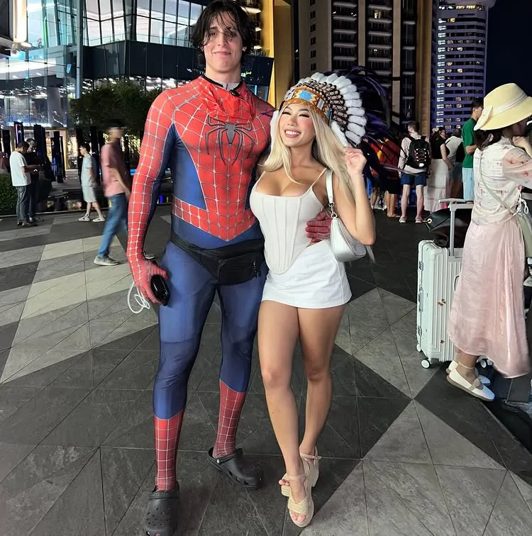 Man dressed as Spider-Man posing with blonde woman in white outfit and feather headdress at a nighttime event in the city