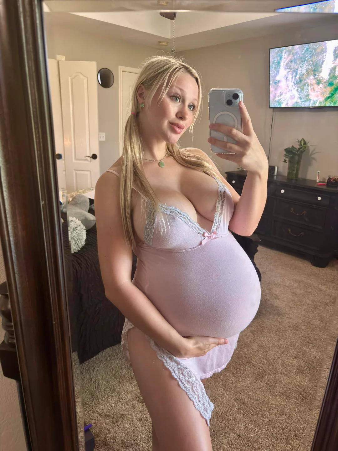 Pregnant blonde woman taking a mirror selfie in a pink lace nightdress, showing her baby bump in a bedroom during late pregnancy