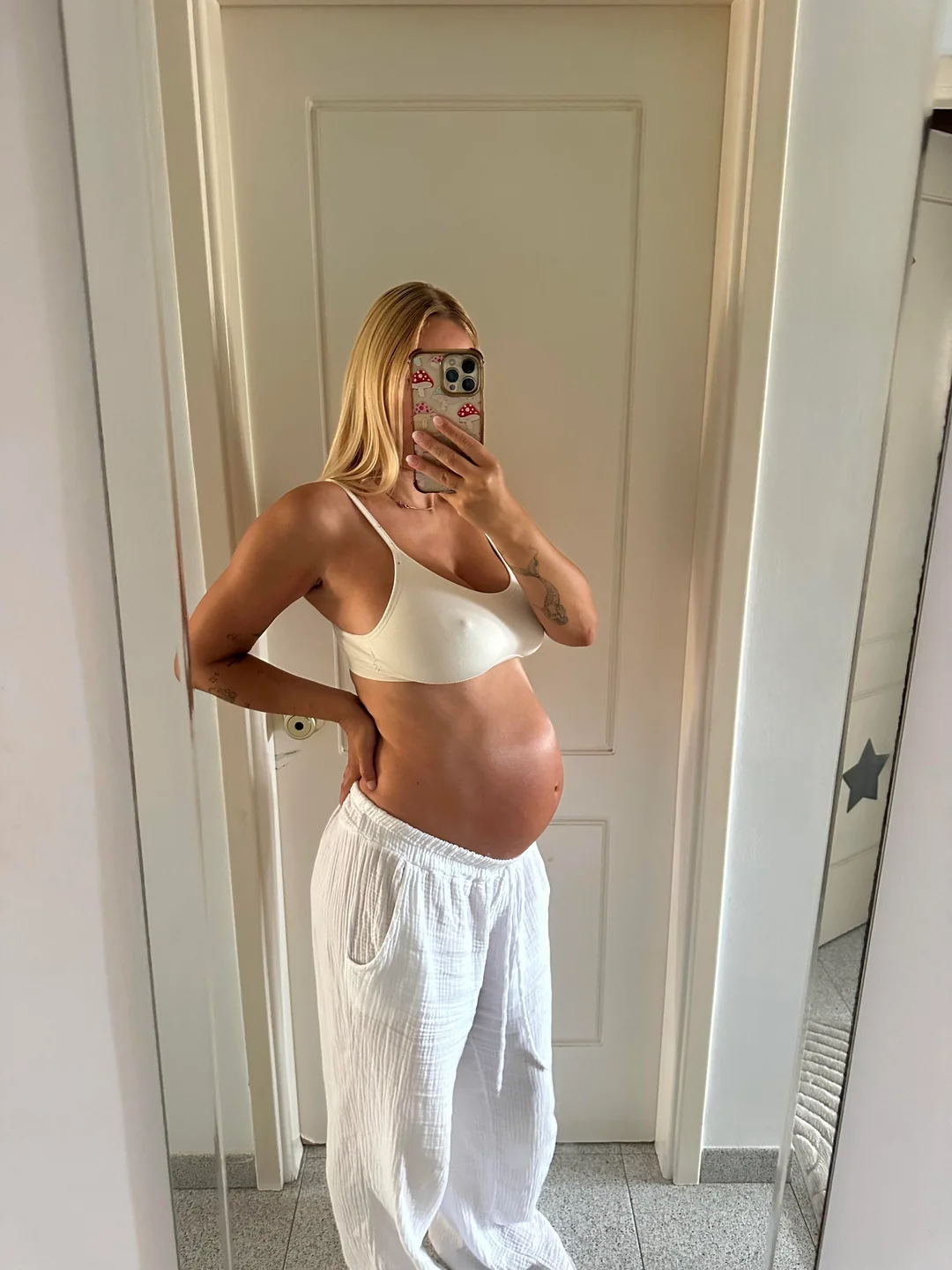 Pregnant Scandinavian blonde woman taking a mirror selfie in a white bra and loose white pants, showing her baby bump indoors
