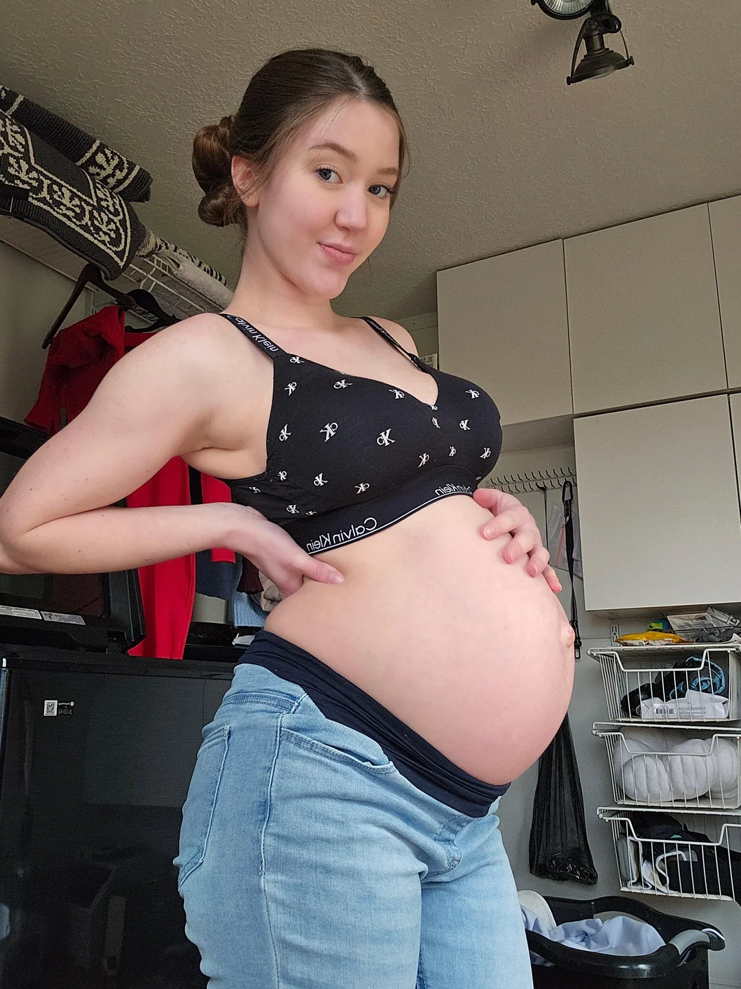 Pregnant brunette woman posing indoors in a black Calvin Klein bralette and jeans, showing her baby bump during late pregnancy