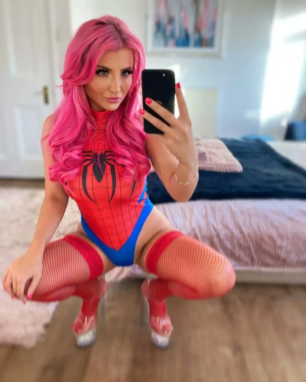 Pink-haired OnlyFans model taking a mirror selfie in a Spider-Man inspired bodysuit with red fishnet stockings and platform heels in a bedroom