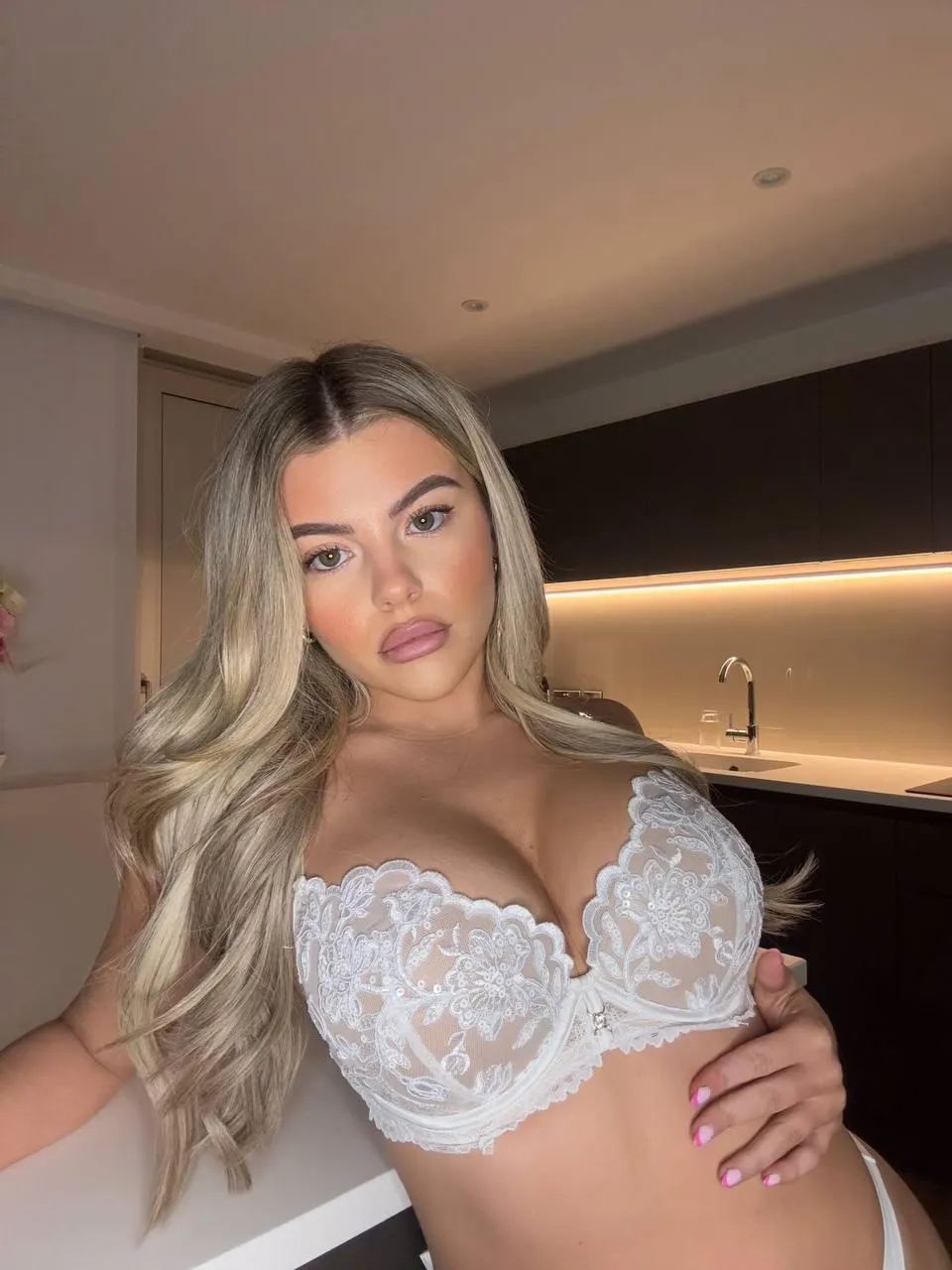 A woman in white lingerie poses confidently in a kitchen, showcasing her style as OnlyFans model Tilly Harrington